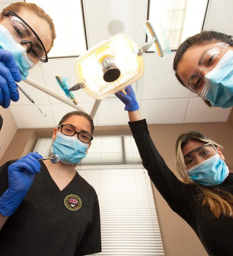 BDG Academy Students looking down from overhead, as viewed from a patient chair. One of the students adjusts the light and another prepares to examine a patient with a dental tool