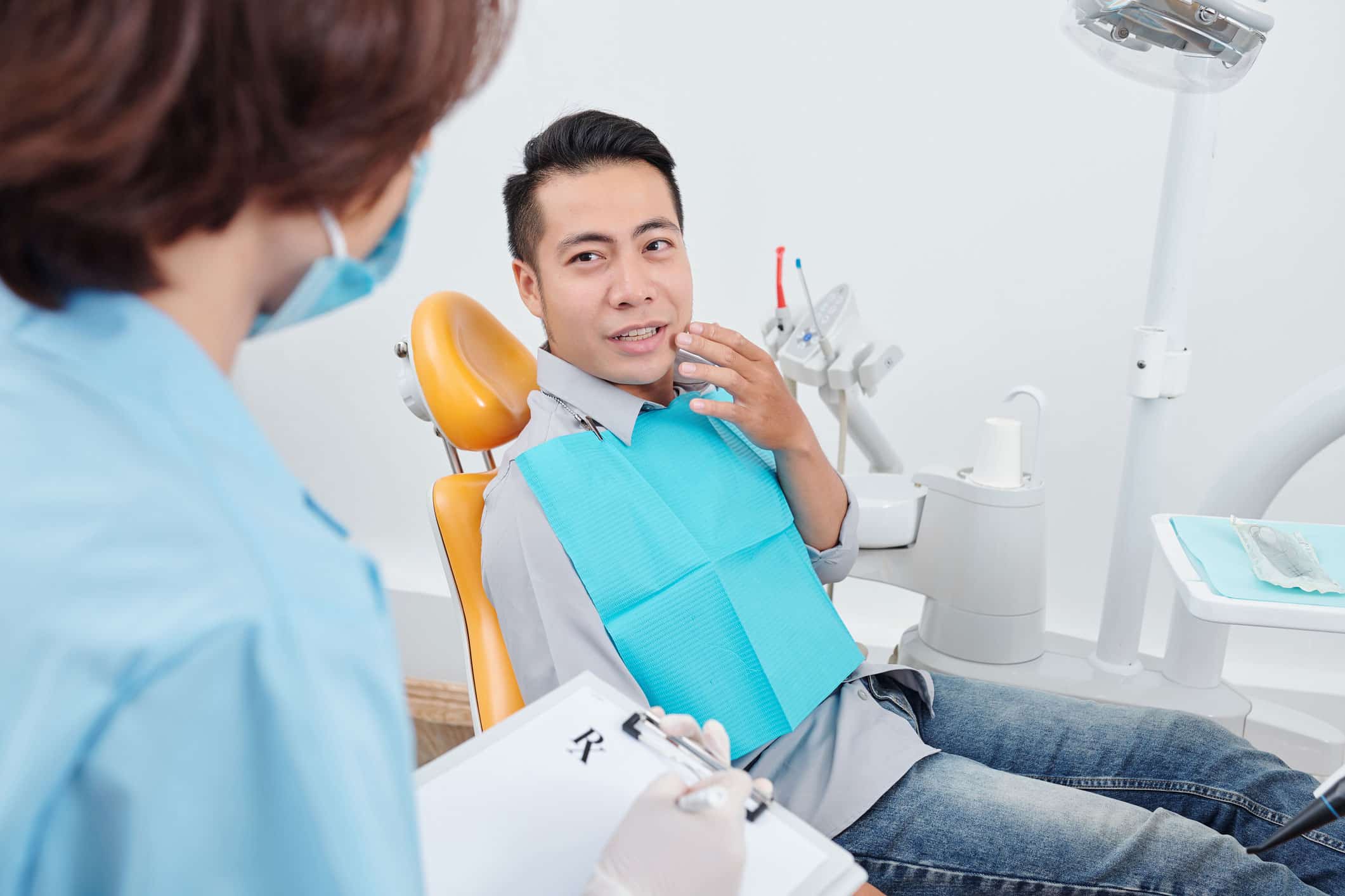 Asian male patient sitting in a dentist office chair lightly touches his jaw, explaining dental pain to a dentist in view, prescribing medication for an infected cavity
