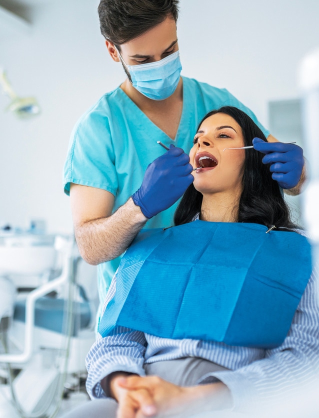 dental patient wearing a blue apron and being examined by a dental hygienist in light blue, standing someone behind her