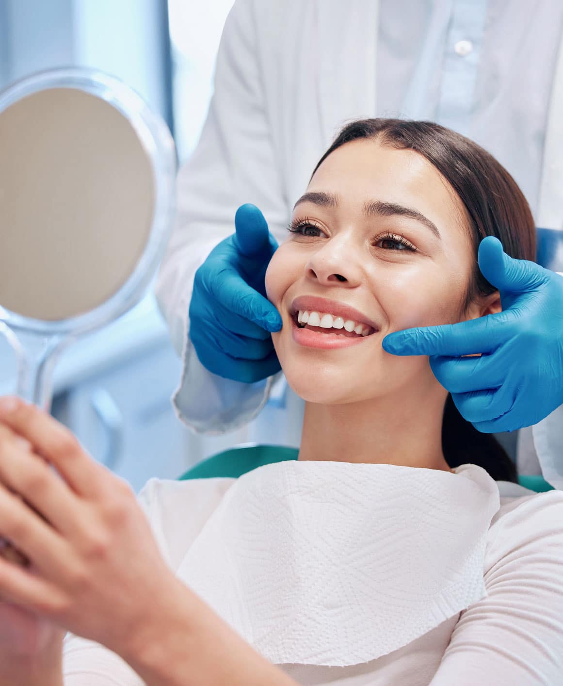 Attractive young female dental patient flashes a straight white smile, looking into a circular mirror as a dentist points to her teeth from behind her, wearing blue surgical gloves