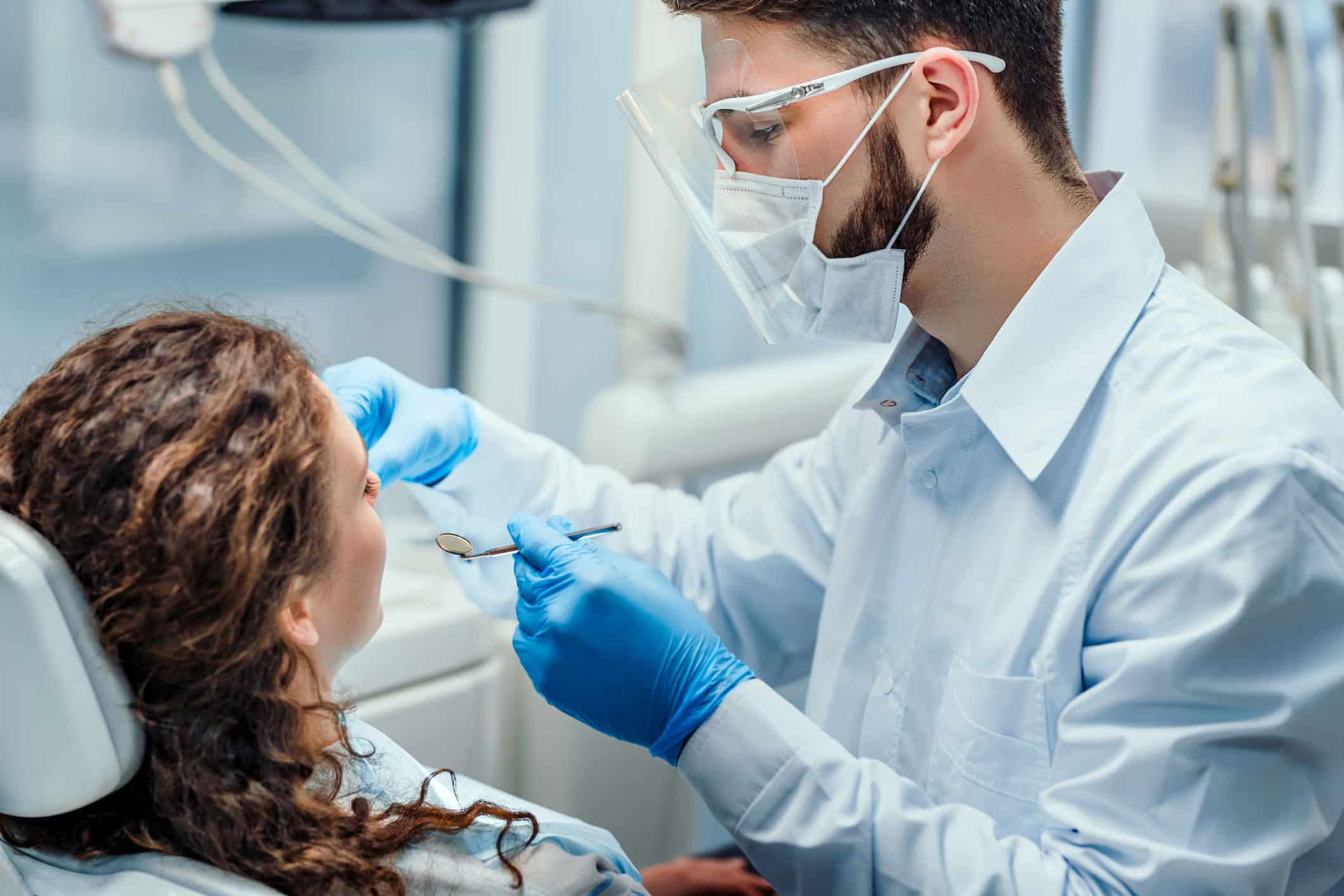 Female patient with curly hair is examined by a male dentist holding dental tools with blue gloved hands.
