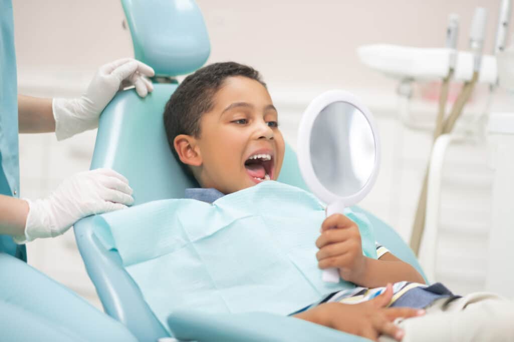 Looking into mirror. Handsome dark-eyed boy opening mouth and looking into mirror while visiting dentist