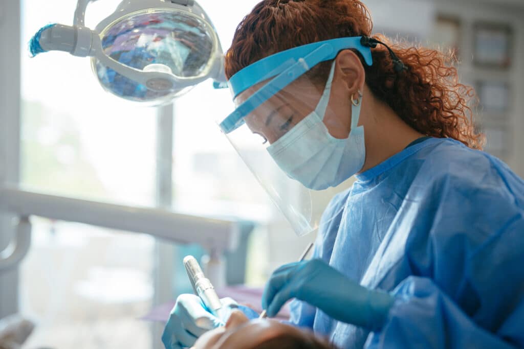 Dentist in blue down and a full plastic face mask performing dental surgery on a patient