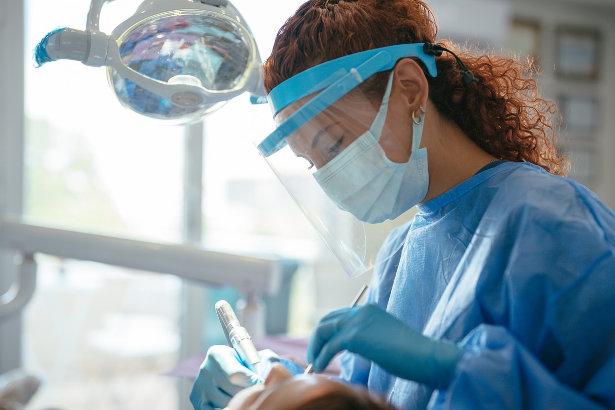 Dentist in blue down and a full plastic face mask performing dental surgery on a patient