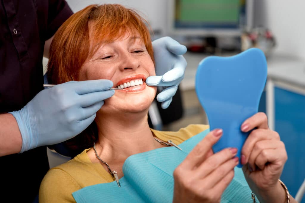Smiling senior woman with new dental implants or dentures sitting in the dental office and looking at the mirror