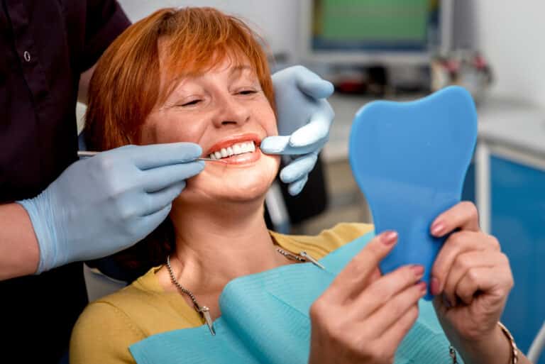 Smiling senior woman with new dental implants or dentures sitting in the dental office and looking at the mirror