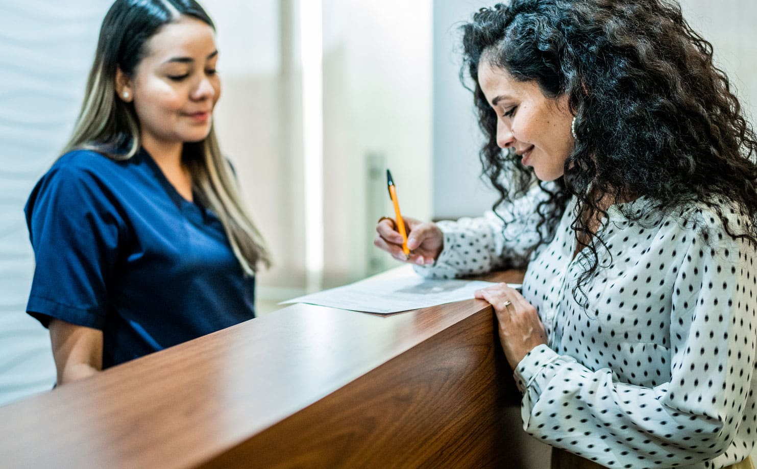 Smiling dental patient filling out insurance forms at a reception desk, while the assistant looks on pleasantly from behind the desk