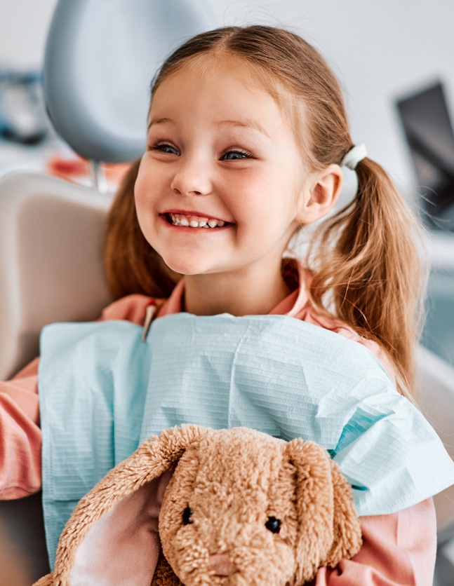 little girl smiled very big from her dentist office chair, a stuffed bunny on her lap