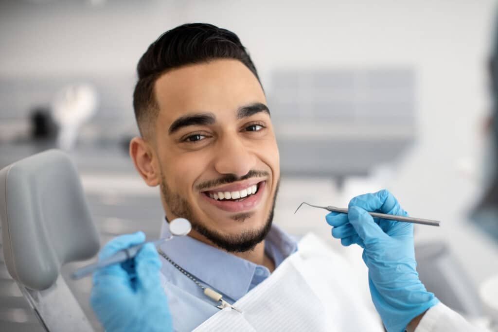 Attractive male dental patient smiles widely from a dentist office chair, two blue gloved hands of a dentist holding dental tools in preparation for an examination