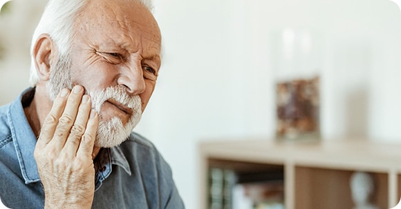 Older senior man sitting at home pushes on his jaw, indicating tooth or jaw pain