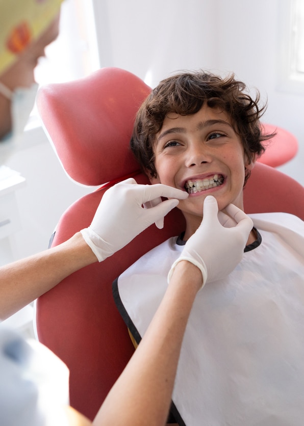 A school-aged ortho patient boy smiles widely while the dental professional checks his retainer