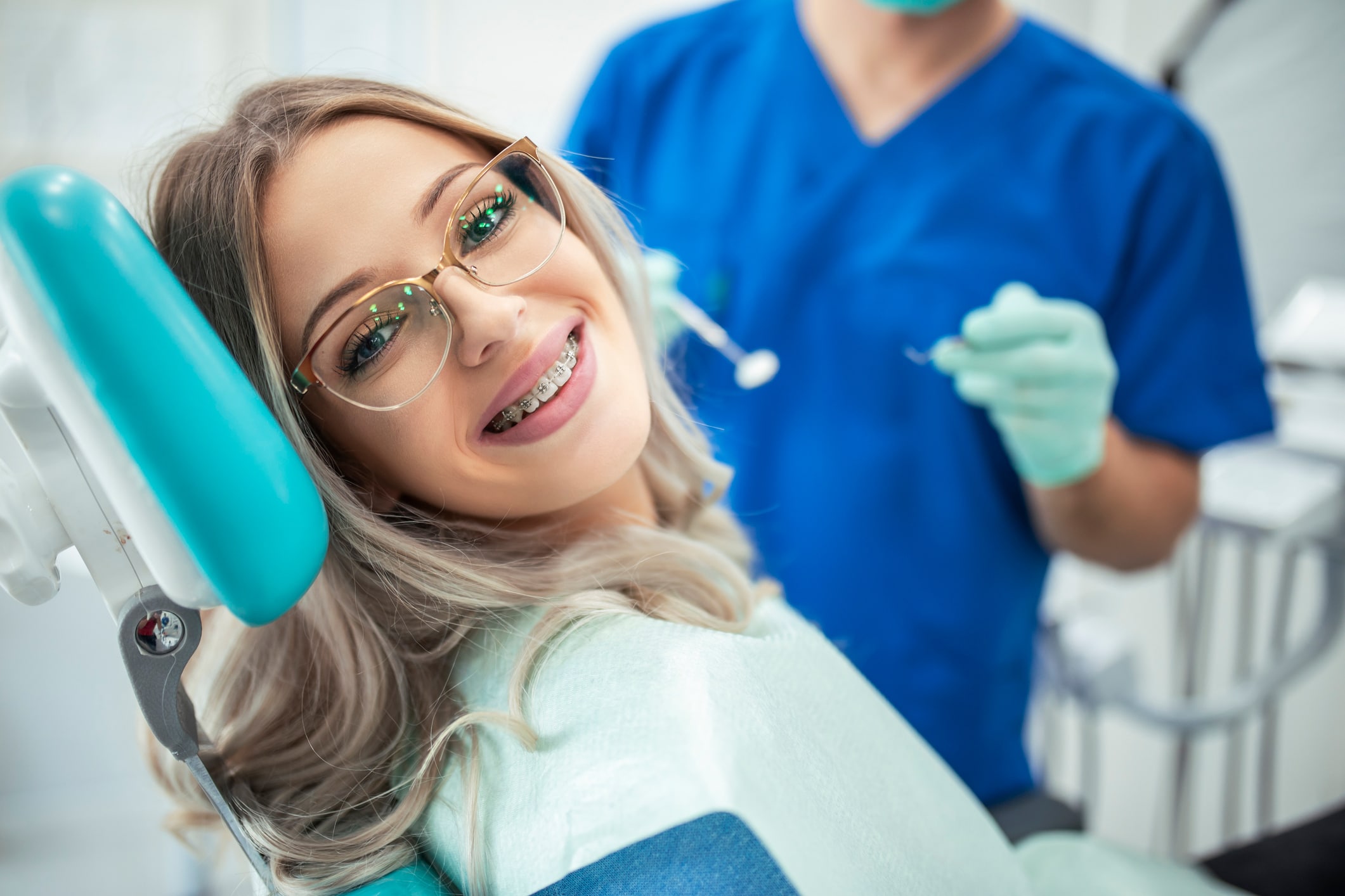 Young female ortho patient wearing glasses smiles from a dental chair, a dentist in blue shown ready for examination in the background