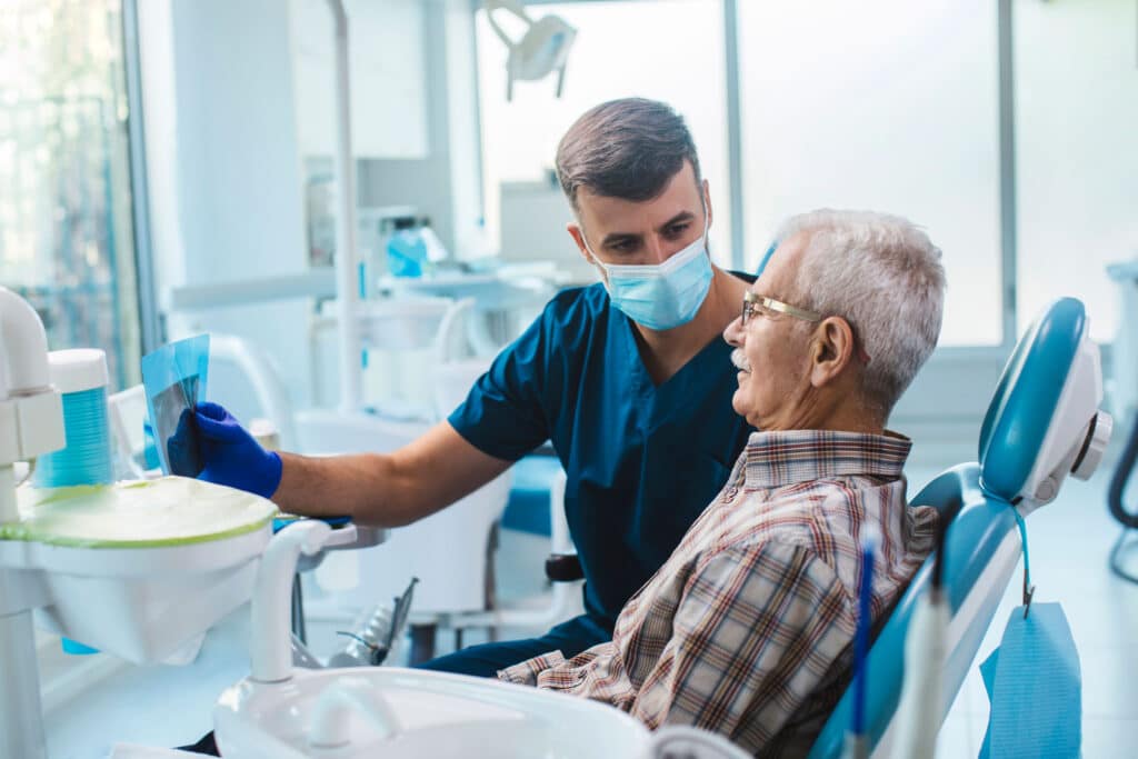 Dentist in dark blue scrubs holds physical x-rays and explains a necessary root canal procedure to a senior male patient wearing a plaid shirt and gold glasses.