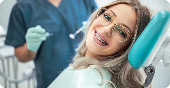 Smiling attractive teenage female ortho patient wearing glasses smiles from a dental chair, a dentist in blue shown ready for her orthodontic examination in the background