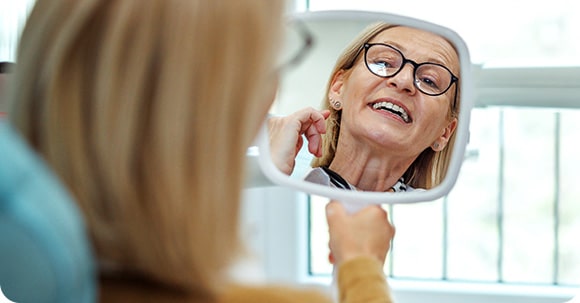 Senior woman with glasses looking at her smile through a square shaped mirror in a dentist office