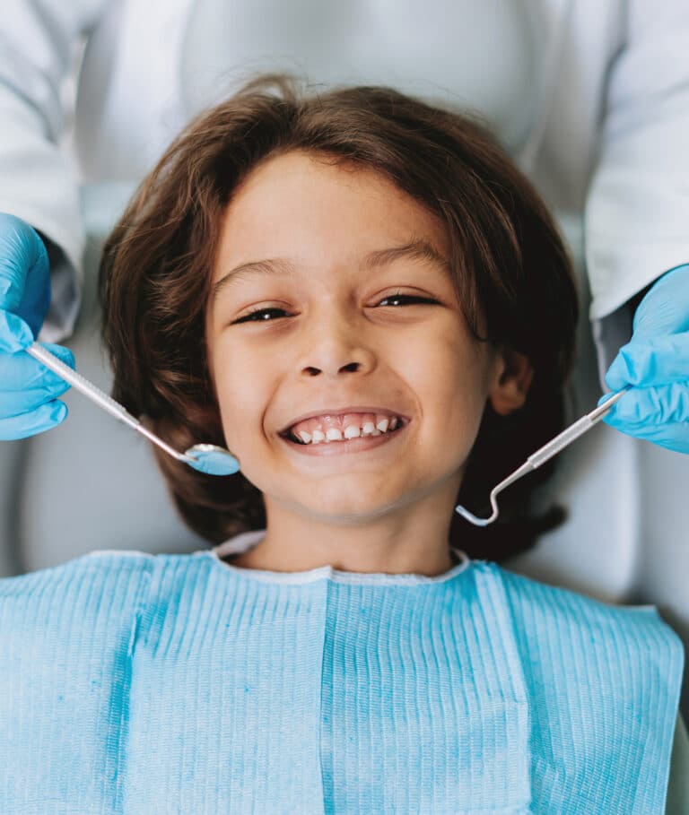 A long haired young boy at the dentist, smiling broadly from the dentist chair, a blue apron across his chest