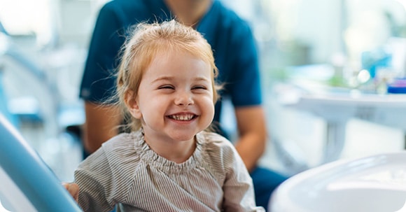 female toddler smiling happily from an examination room chair, with her dentist in view, blurred in the background